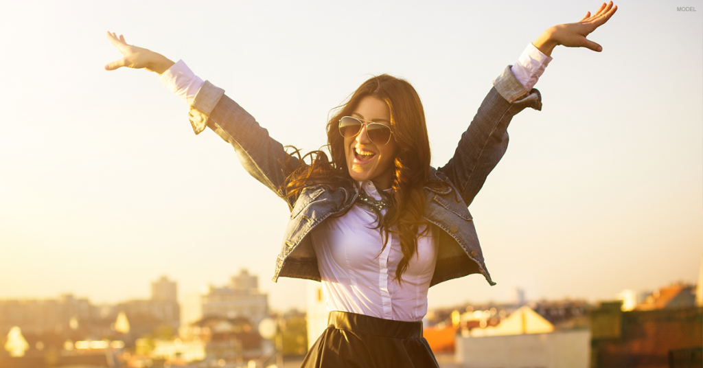 Young attractive woman in sunglasses smiling with both arms raised above her head.