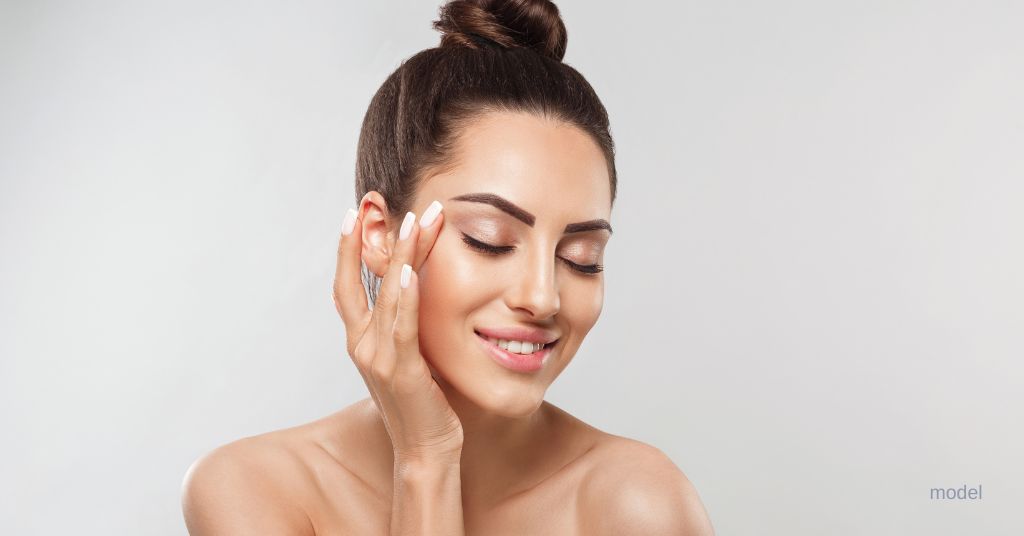 Beautiful brunette woman smiles while looking down and holding her face. (Model)